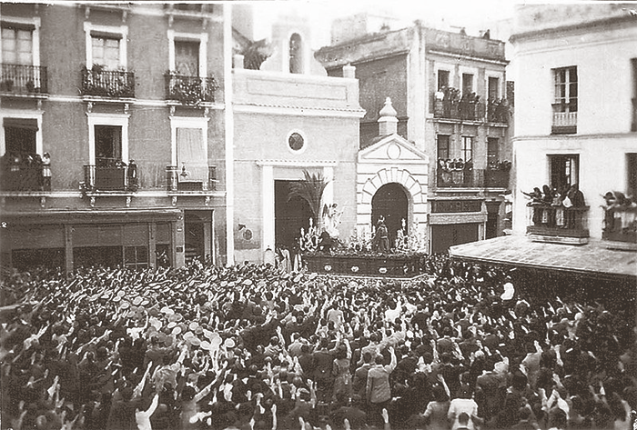 Jueves Santo de 1937: Sobre el paso del Cristo de la Buena Muerte prestado para la ocasión por la Hermandad de la Hiniesta y con una palmera en el mismo, vemos saliendo de su templo al Señor de la Oración en el Huerto de la Hermandad de Montensión, en tanto es saludado brazo el alto por los cientos de personas congregadas en la Plaza de los Carros presenciando su salida cofradíera.